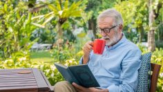 Senior elderly man reading book drinking mug of coffee in garden